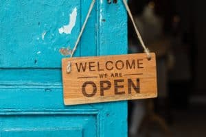 Wood sign hanging off of a door knob that says "welcome, we are open"